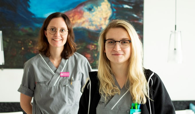 Två kvinnor står framför en stor målning och ler in i kameran. De har båda på sig en grå skjorta och glasögon.