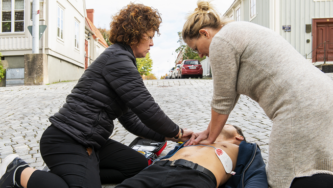 Två kvinnor gör hjärt-och lungräddning på en person som ligger på rygg på marken. Personerna är utomhus, på en kullerstensbelagd gata.