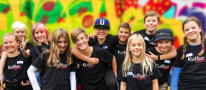 Glada ungdomar iklädda svarta KulturCrew-tröjor framför en färgglad grafittibakgrund.