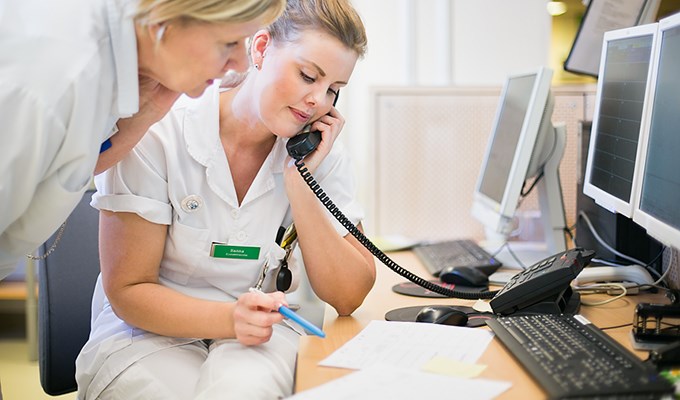 En sjuksköterska sitter och pratar i telefon vid ett skrivbord och läser samtidigt på ett papper. En kollega lutar sig fram bredvid och läser på samma papper.