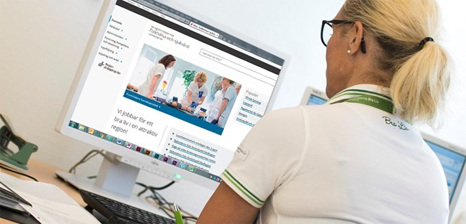 En vårdklädd kvinnlig personal sitter framför en datorskärm. Datorskärmen visar webbplatsen "Folkhälsa och sjukvård i Jönköpings län"