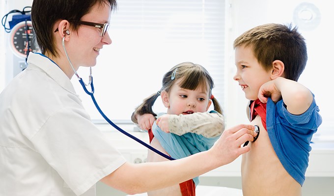 En läkare med stetoskop i öronen och två barn som dragit upp sina tröjor så att läkaren kan lyssna på deras hjärta.