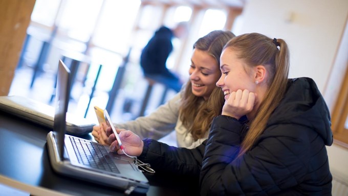 Två leende tjejer som tittar i sina smartphones samtidigt som de arbetar vid en laptop.