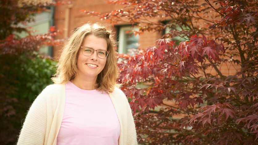 Anna står utomhus framför en japansk lönn. Hon ler, har glasögon, blont hår och en vit kosta på sig.
