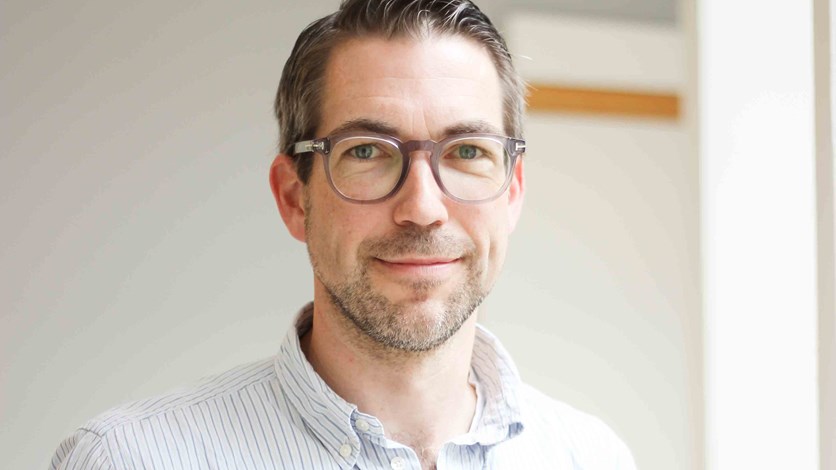 Michael har mörkt lite gråsprängt hår och glasögon. Han har en ljus skjorta och står i en inomhusmiljö. Han ser glad ut.