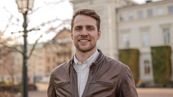 Simon har en brun skinnjacka och står utomhus i fint höstväder.