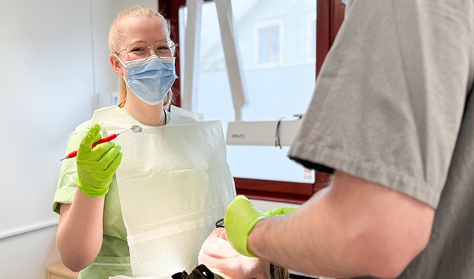 En tandläkare behandlar en patient som ligger ner i en behandlingsstol. Tandläkaren kollar in i kameran och ler.