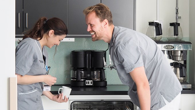 Två personer, en man och en kvinna, står i ett kök framför en diskmaskin. Båda böjer sig lite framåt och skrattar.