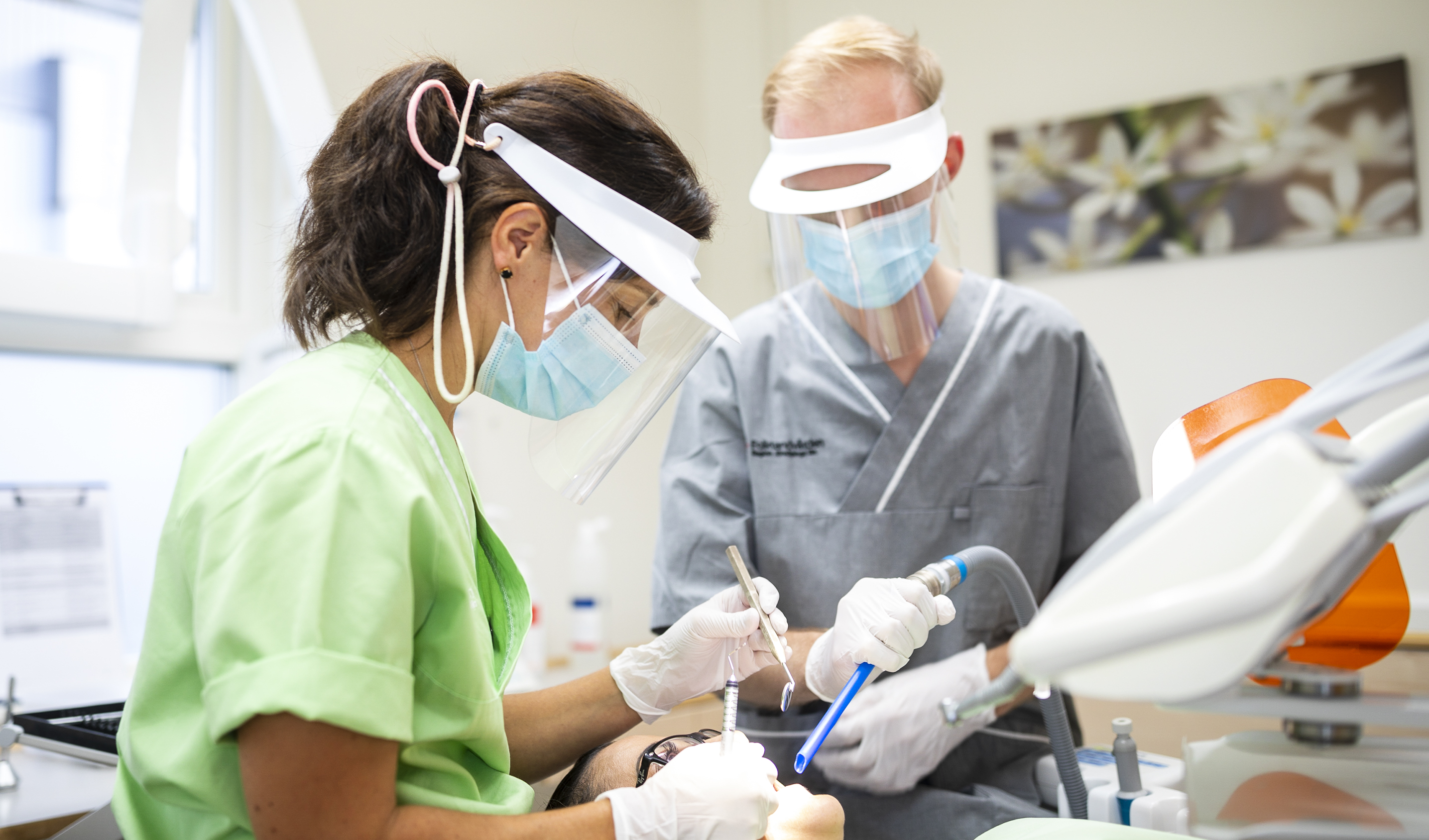 Två personer, en man och en kvinna, i ett behandlingsrum. De utför en behandling på en patient som ligger i tandläkarstolen. Båda behandlarna har visir på sig och tittar ner på patienten.
