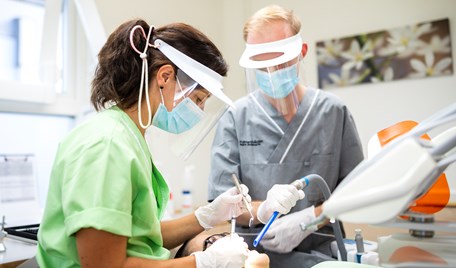 En kvinna och en man står i en klinisk tandvårdsmiljö och behandlar en patient. Kvinnan har brunt hår och grön tröja. Mannen har blont hår och grå tröja. De står börja över patienten med skyddsvisir, munskydd och vita handskar.