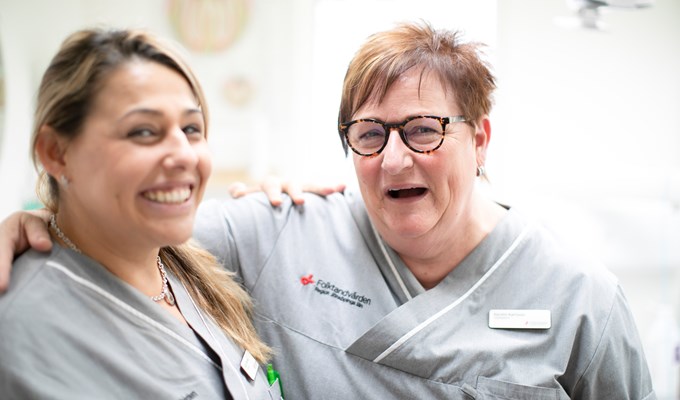 Två kvinnor i klinikkläder håller om varandra och skrattar in i kameran. 