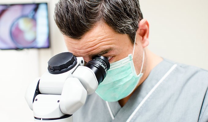En man med munskydd tittar ner i ett mikroskop. 