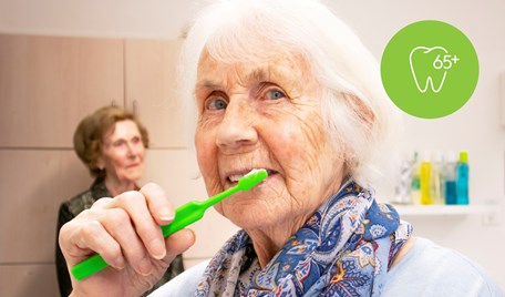 Äldre kvinna borstar tänderna och ler in i kameran. Hon står i ett behandlingsrum. Bakom henne står en annan äldre kvinna.