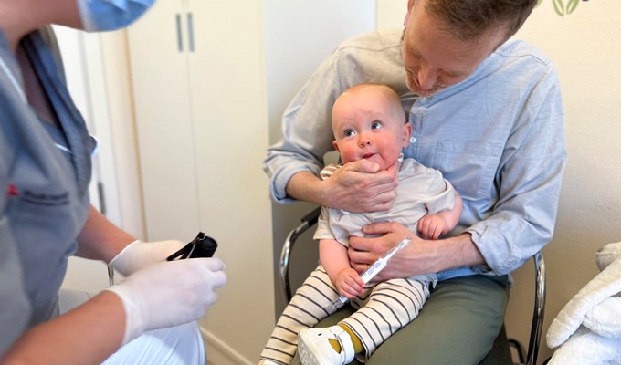 Liten kille sitter i pappas knä och ler mot tandsköterskan som sitter mitt emot och vill att han öppnar munnen.