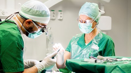 Två personer står böjd över en patient under operation. De båda har gröna kläder, munskydd och skyddsmössa.