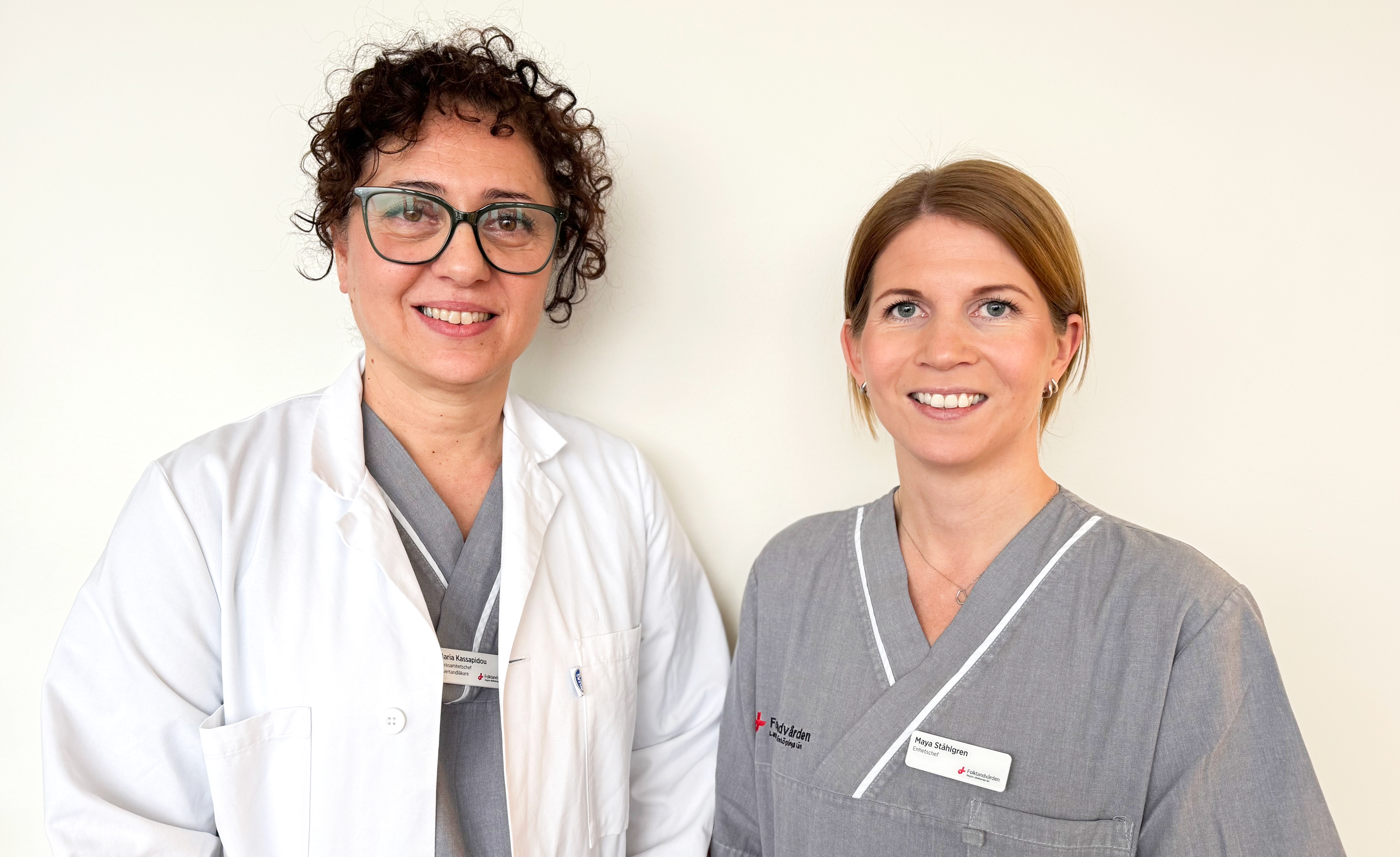 Två kvinnor står i klinikkläder och ler in i kameran.