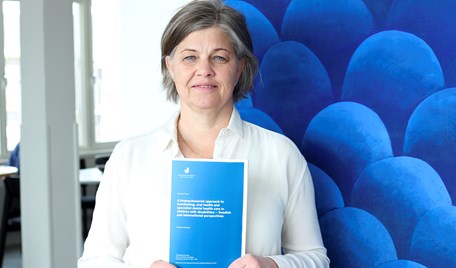 En kvinna står och håller i en blå bok framför en blå tavla.