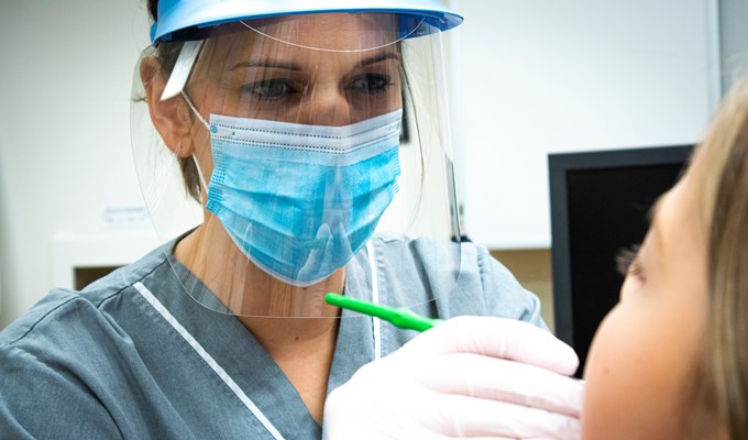 Kvinna med visir och munskydd tittar i munnen på en barnpatient. Hon har grå tröja och vita handskar. 