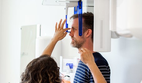  En vuxen patient står i en röntgenmaskin. En tandsköterska rättar till maskinen.