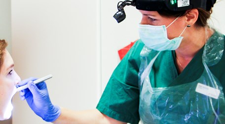 En kvinna med lyser med en ficklampa i en patients mun. Hon har munskydd, grön tröja och en pannlampa.
