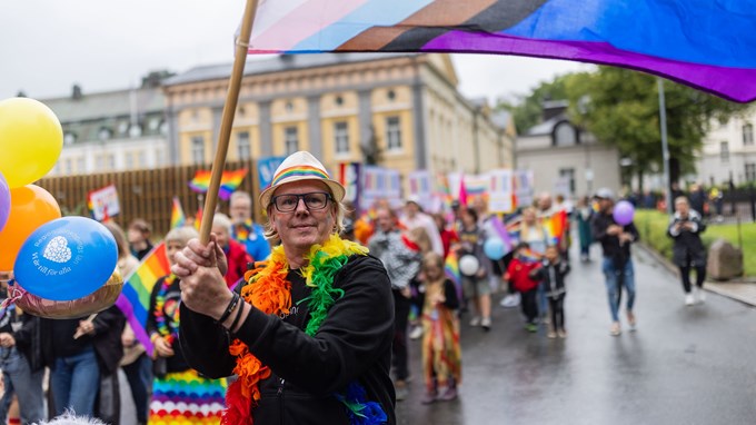 Man med färgglad utstyrsel som viftar med en regnbågsflagga med en prideparad i bakgrunden