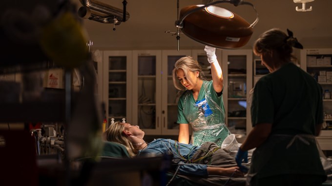 En patient ligger på en säng med en stark lampa över sig. Två medarbetare på akutkliniken står bredvid sängen och tar hand om patienten.