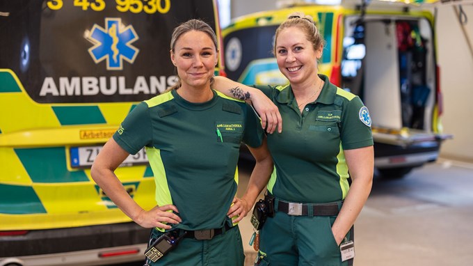 Två medarbetare står bredvid varandra och ler mot kameran. De har klädda i arbetskläder för ambulanspersonal