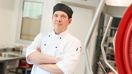 En person ler mot kameran och han har kock-kläder på sig. Han står i ett kök på sin arbetsplats