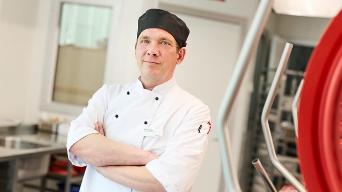 En person ler in mot kameran och han har på sig kock-kläder. Han står i ett stort kök på sin arbetsplats. 