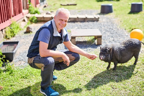 En person sitter på huk och ler mot kameran och klappar en mini-gris. 