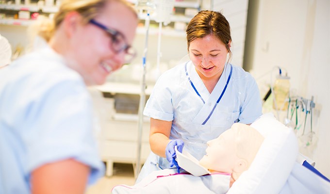 Två kvinnor i ljusblå sjuksköterskekläder undersöker dockan som används som fiktiv patient. 