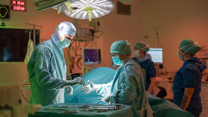 Två medarbetare jobbar i en operationssal och de har operationskläder på sig. Den ena personer ger ett verktyg till den andra personen. 