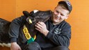 Person i arbetskläder sitter med en kalv som slickar honom i ansiktet, båda ser glada ut framför en orange bakgrund.