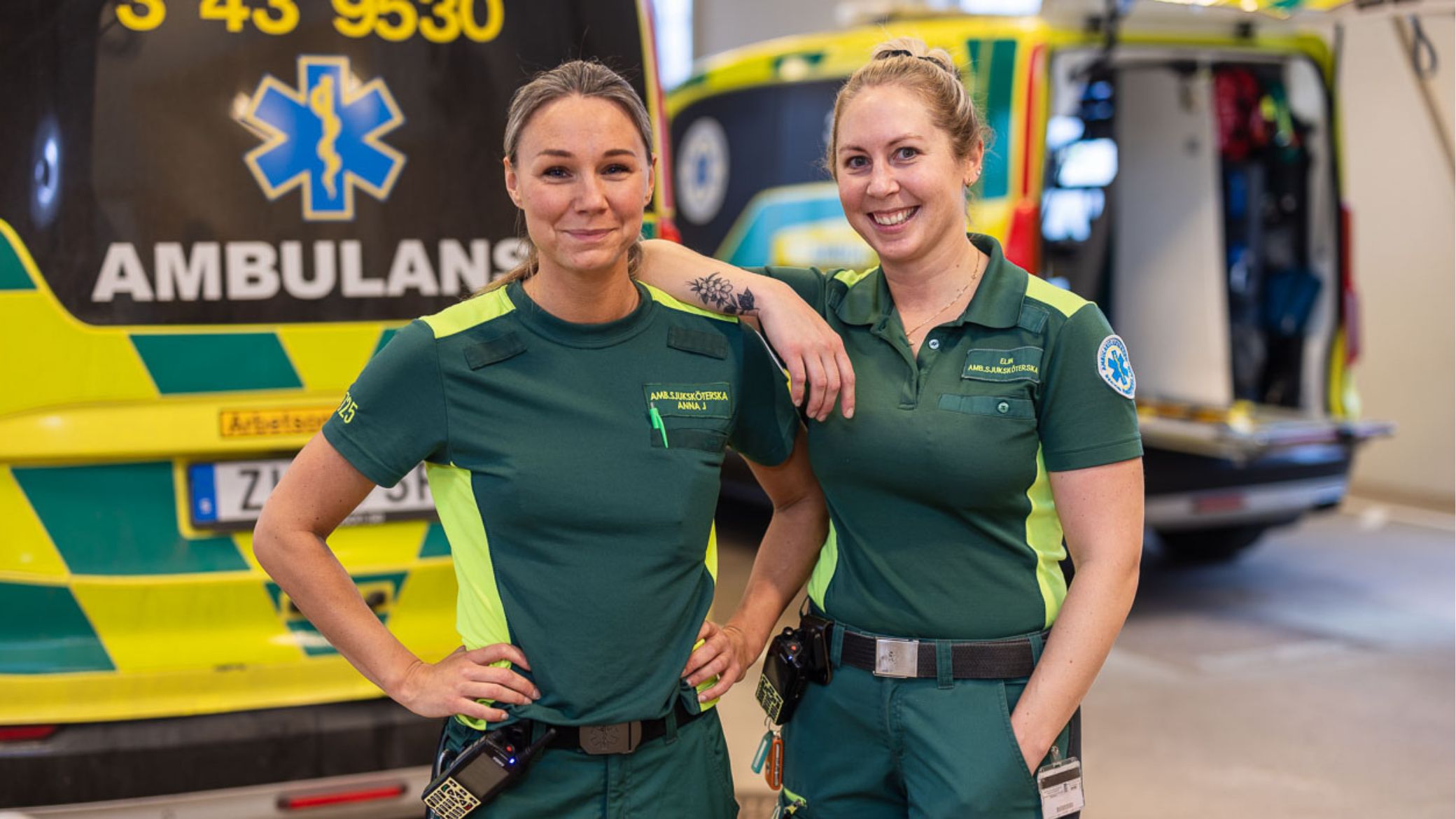 Två kvinnliga ambulanssjukvårdare står framför en ambulans. De tittar rakt fram mot betraktaren och ler. Den ena lutar armen mot den andra kvinnan.