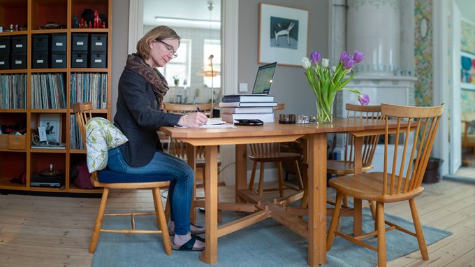 En kvinna vid ett köksbord antecknar i ett block. Tulpaner i en vas på bordet. En laptop står på fyra böcker, för att få bildskärmen i rätt höjd. 