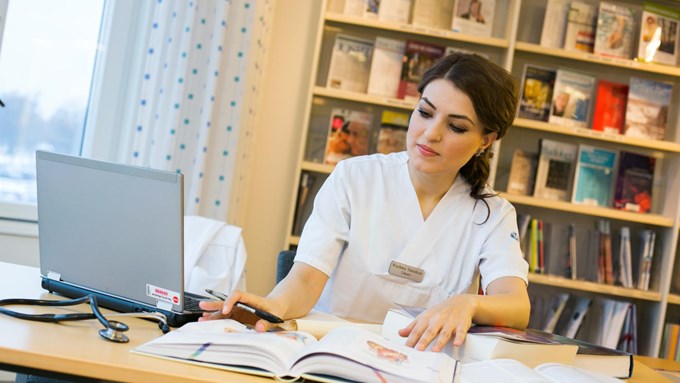 Läkaren Wazhma Stanikzai sitter vid ett bord med dator och medicinsk bok framför sig och har en penna i handen. Befinner sig i biblioteksmiljö.