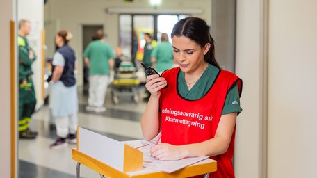 Kvinna med gröna sjukvårdskläder och en röd väst med texten ledningsansvarig ssk akutmottagning står vid en högt bord och håller upp en walkie talkie