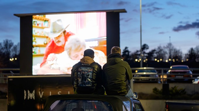 Två ungdomar sitter på taket till sin bil och kollar på utomhusbio på en parkering.  