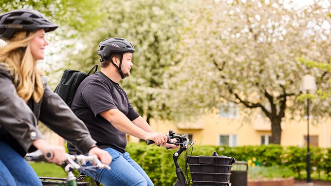 E man och en kvinna cyklar bredvid varandra. I bakgrunden blommande träd i en stadspark. 