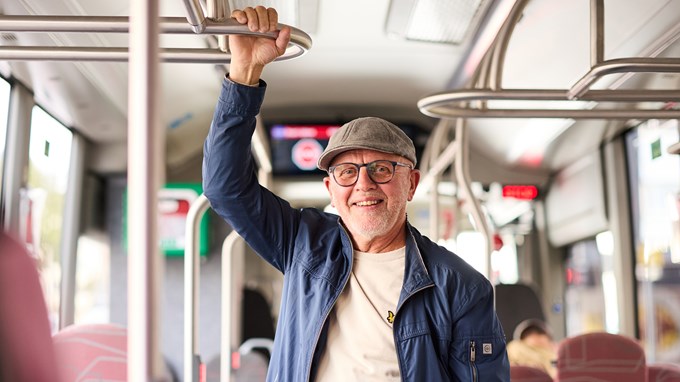 En äldre man med keps står i en buss och håller sig i ett handtag i taket. 