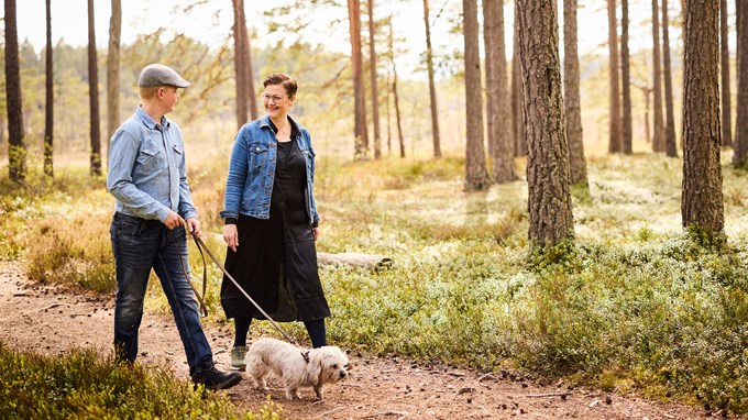 En ung man och kvinna är ute och rastar hunden på en stig med blåbärsris runt omkring. 