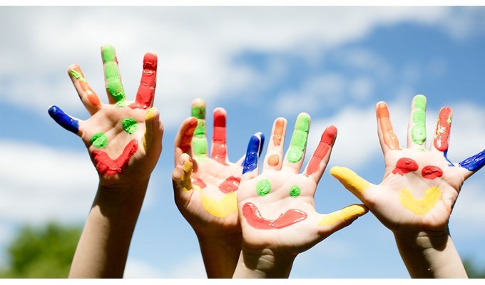 Fyra barnhänder sträcks upp mot en blå himmel. På händerna är det målat glada ansikten i gult, grönt, rött och blått. 