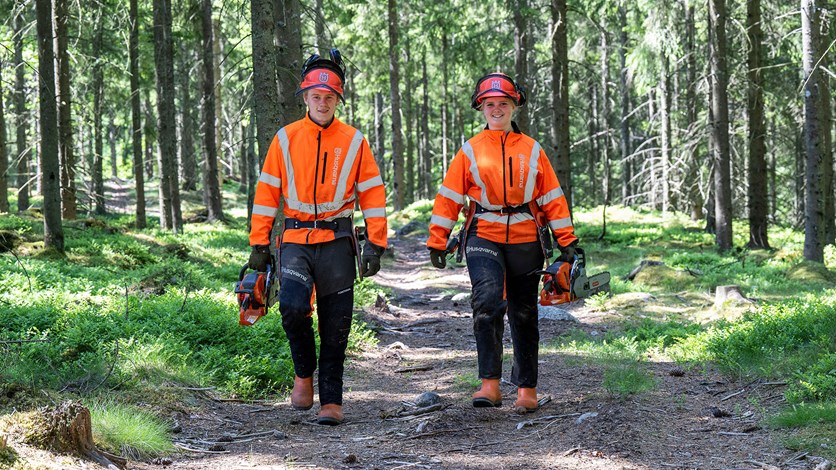 Två elever med orangea varselkläder går i en ljus grönskande skog med motorsågar i handen.
