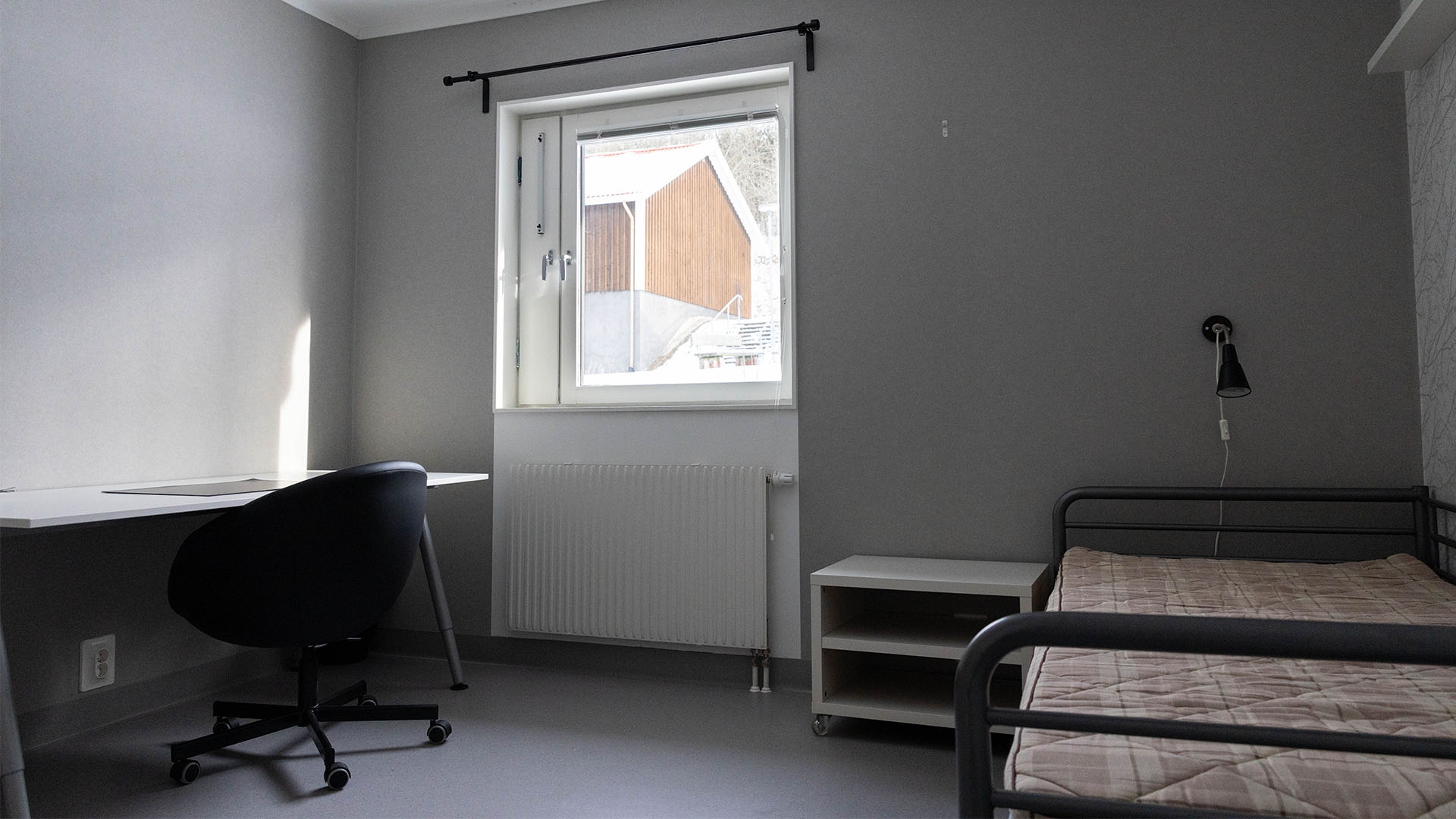 Ett sovrum med grå väggar, en säng, sängbord, lampa och ett skrivbord med stol.