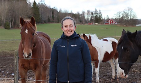Josefine står utanför en hästhage med två bruna hästar i bakgrunden