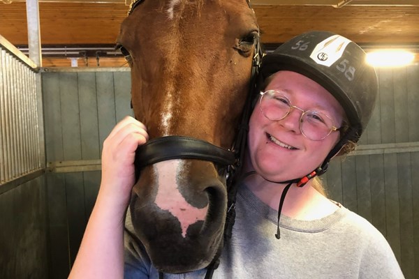 Glad kvinna med glasögon och ridhjälm håller i en brun häst