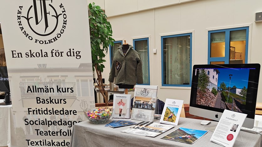 Bord med grå duk. På bordet står utbildningskataloger och foldrar. Till höger en bildskärm som visar en bild från Värnamo Folkhögskola.